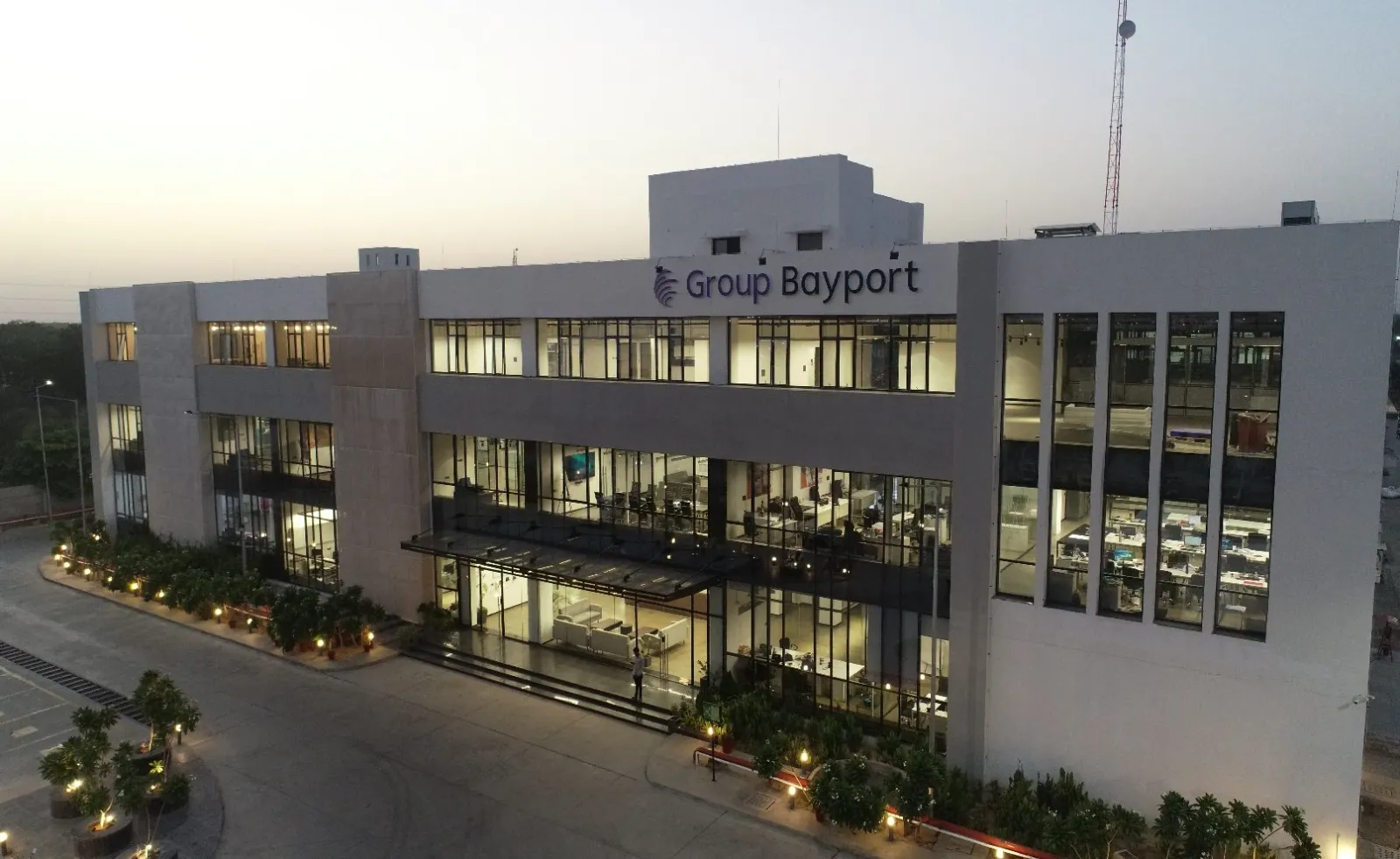 Drone view at dusk of a lit three-storey glass office building with landscaped driveway lights