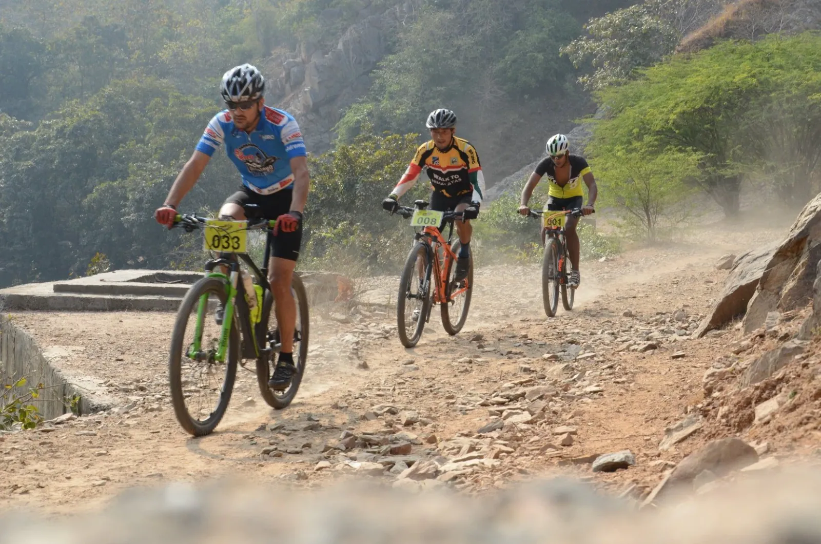 Three mountain bikers riding a dusty rocky trail through green hills during a race