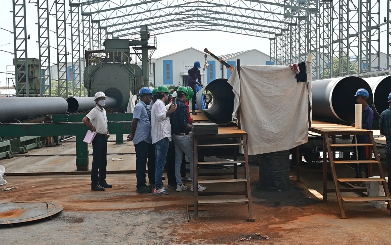 Engineers in hard hats inspect large coated steel pipes inside an open steel-frame industrial shed