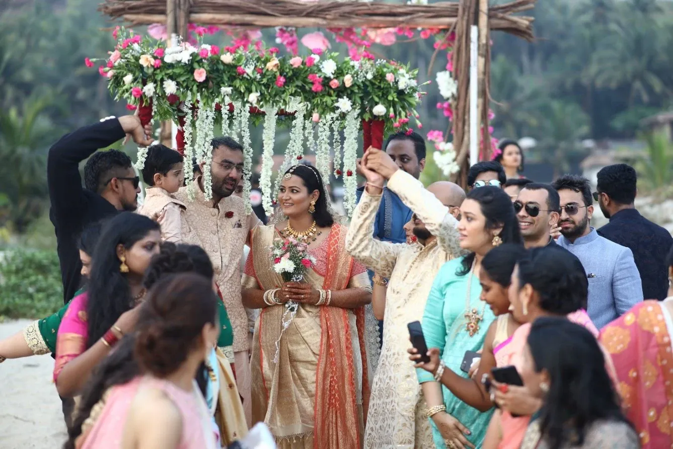 Bride in a gold saree walks under a floral canopy held by family at a beachside wedding