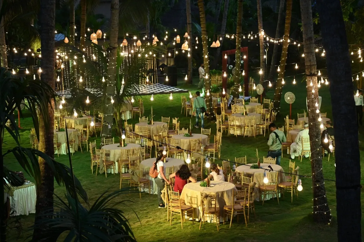 Outdoor evening event with round tables, gold chairs and string lights among palm trees